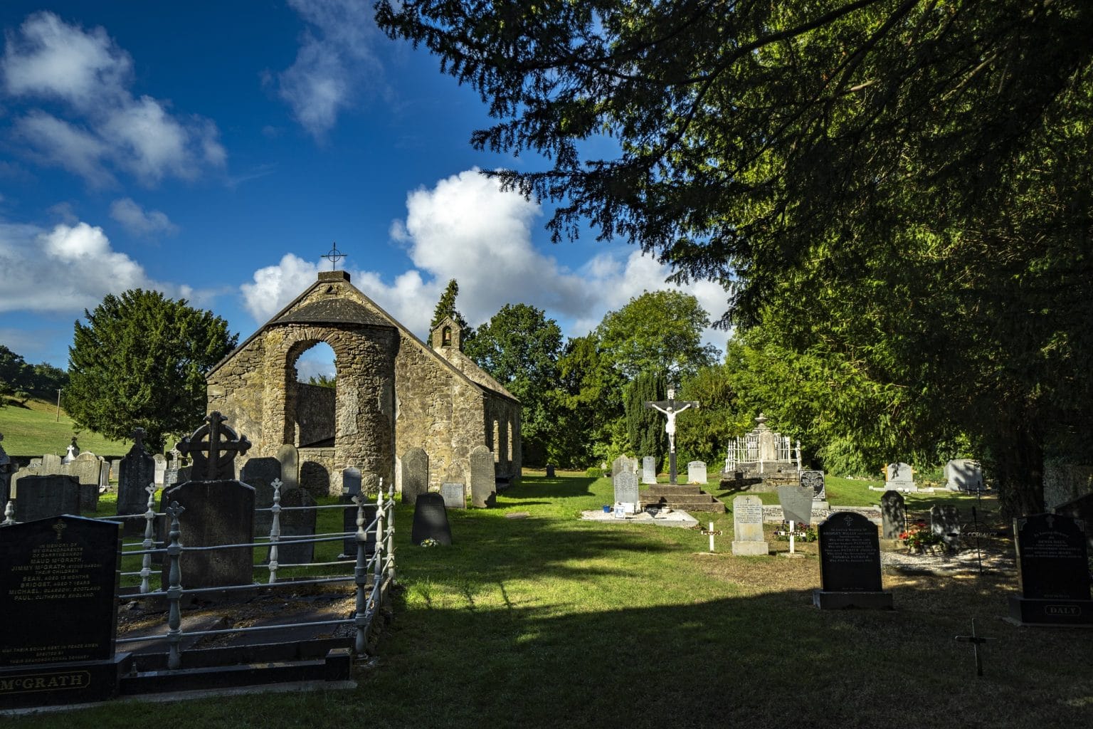 Grassy cemetery with various gravestones and a weathered stone chapel with an arched entrance and cross on top, set against a bright blue sky with scattered clouds and surrounded by green trees.