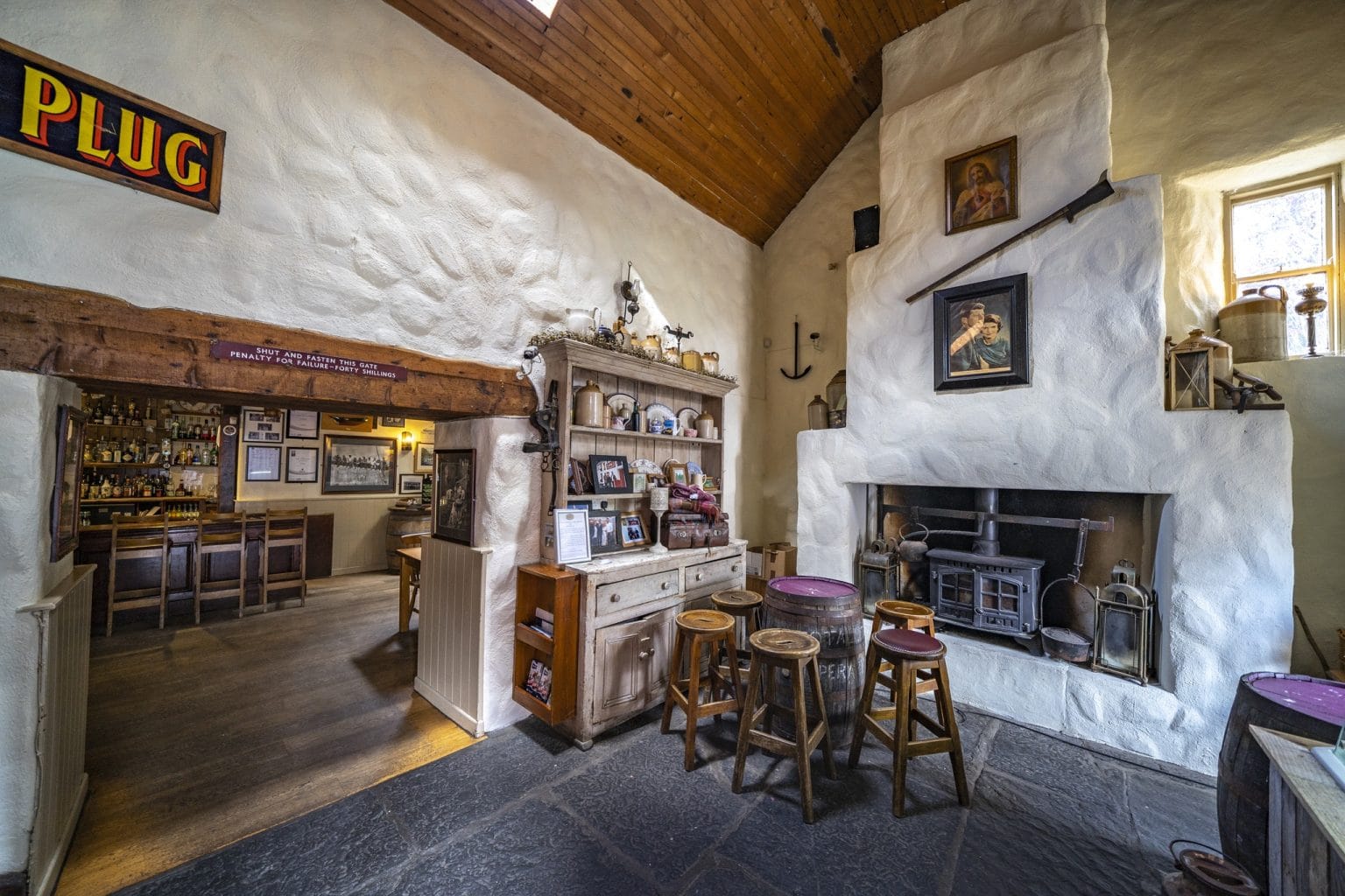 Cozy rustic pub interior featuring white textured walls and wooden ceiling, a small sitting area with stools around a barrel table, an old fireplace with framed pictures above, shelves filled with vintage pottery and photos, and a bar visible through a wooden beam doorway with a sign reading "PLUG."