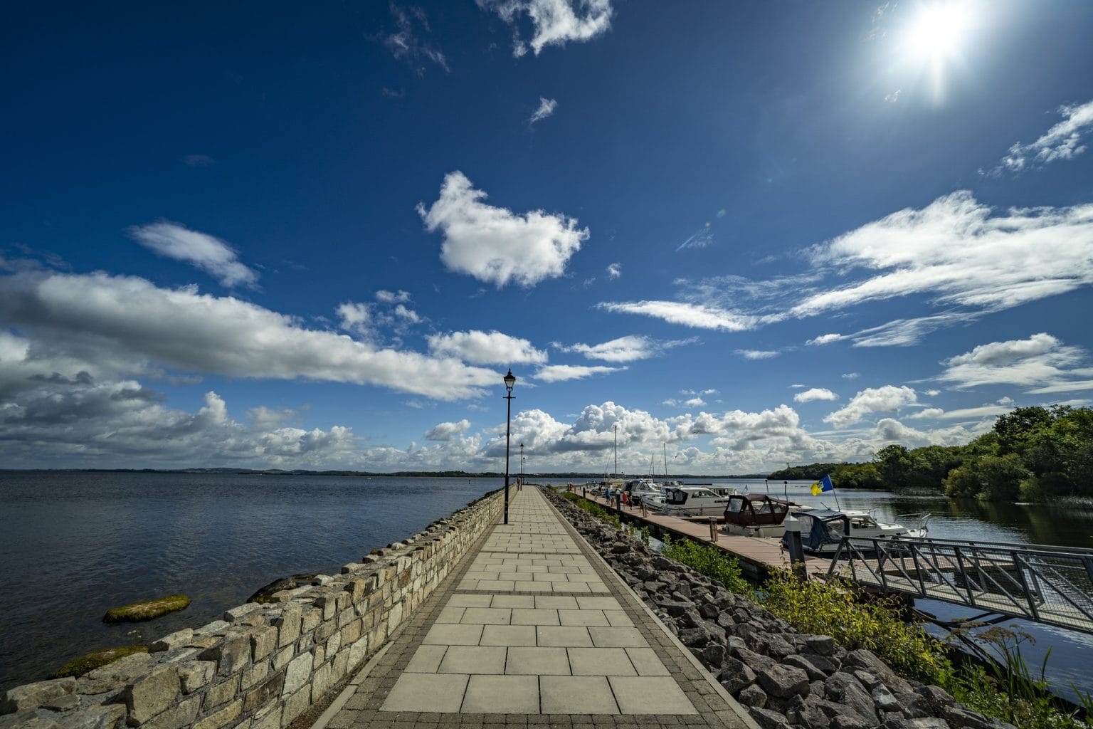 Stone-paved pier extending into a large body of water under a bright blue sky with scattered clouds, with boats docked on the right side and a low stone wall on the left.