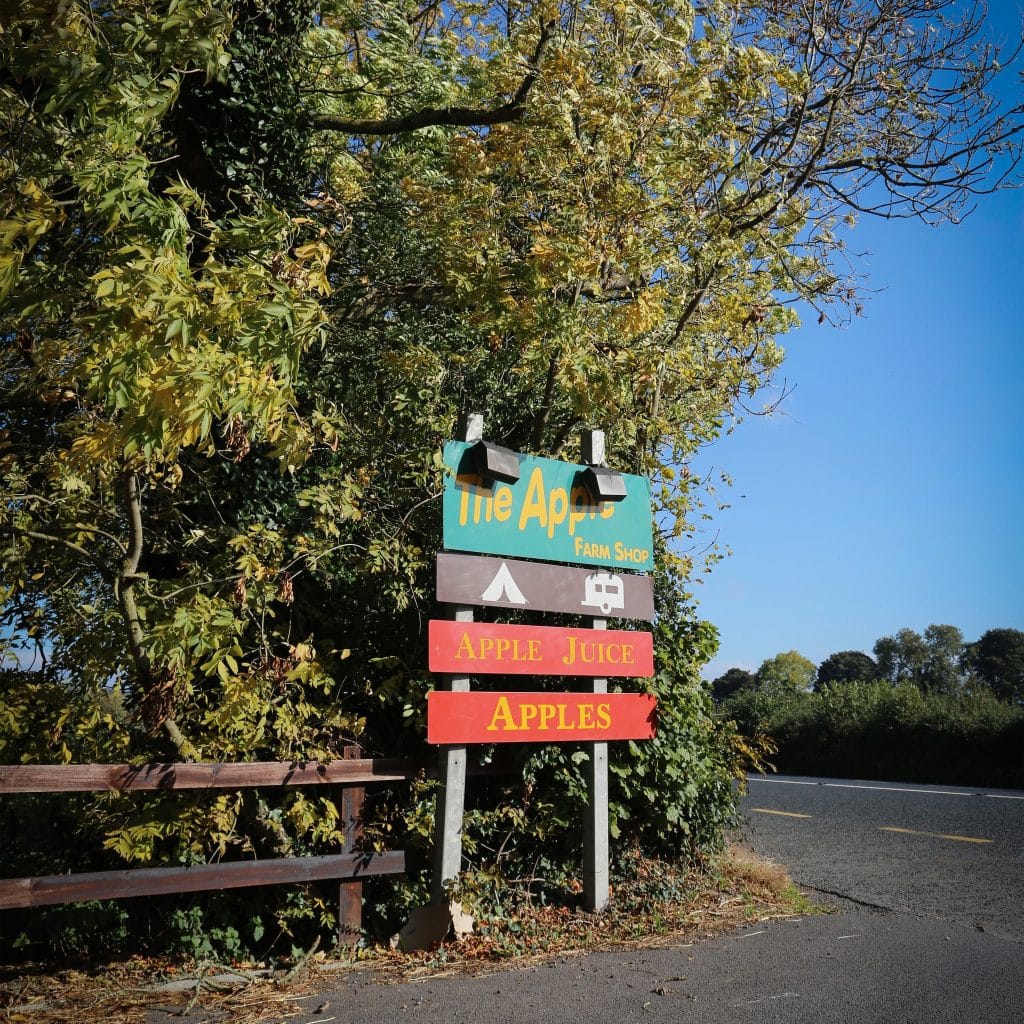 Roadside sign for "The Apple Farm Shop" with additional signs for apple juice and apples, set against green trees and a clear blue sky.