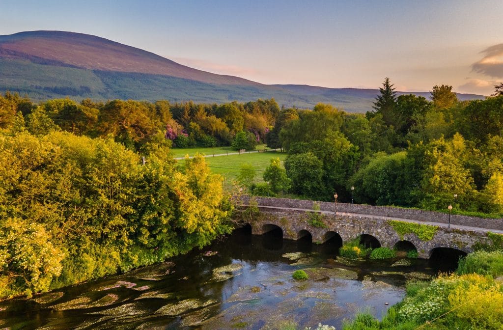 Stone arch bridge over a calm river surrounded by lush green trees and vegetation, with a mountain and clear sky in the background during sunset.