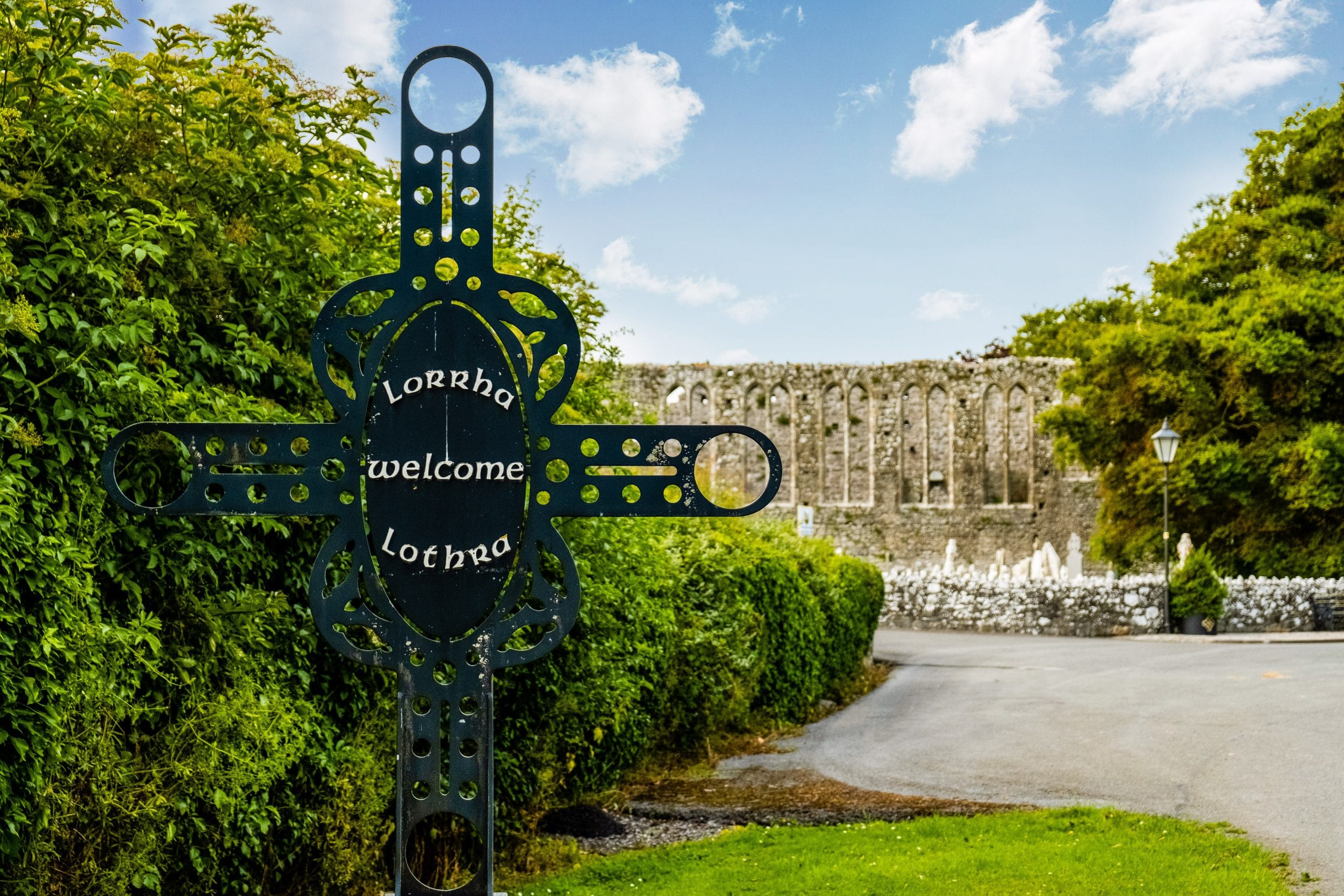 A black metal cross-shaped welcome sign with the words "Lorrha welcome Lothra" stands next to a green hedge along a curved road, with ancient stone ruins and a bright blue sky with scattered clouds in the background.
