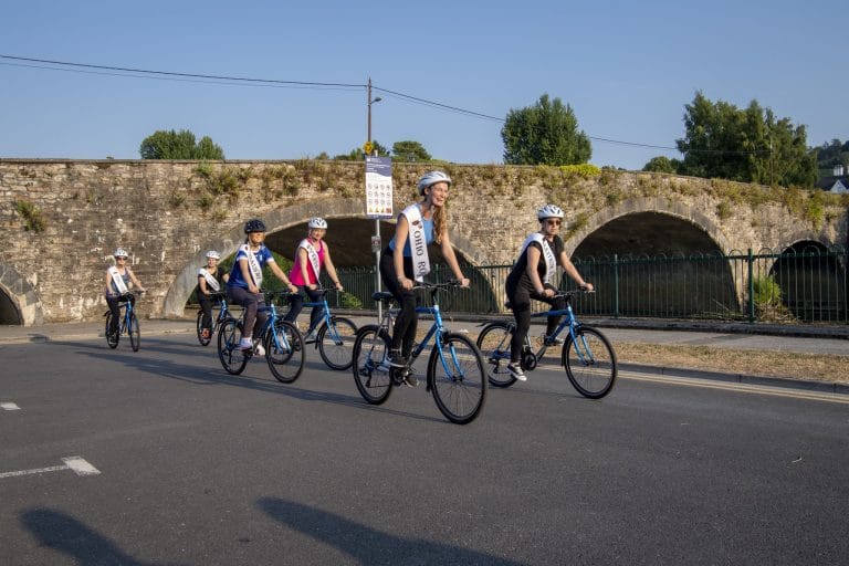 A group of six women wearing helmets and sashes riding blue bicycles on a paved road, with an old stone bridge and clear blue sky in the background.