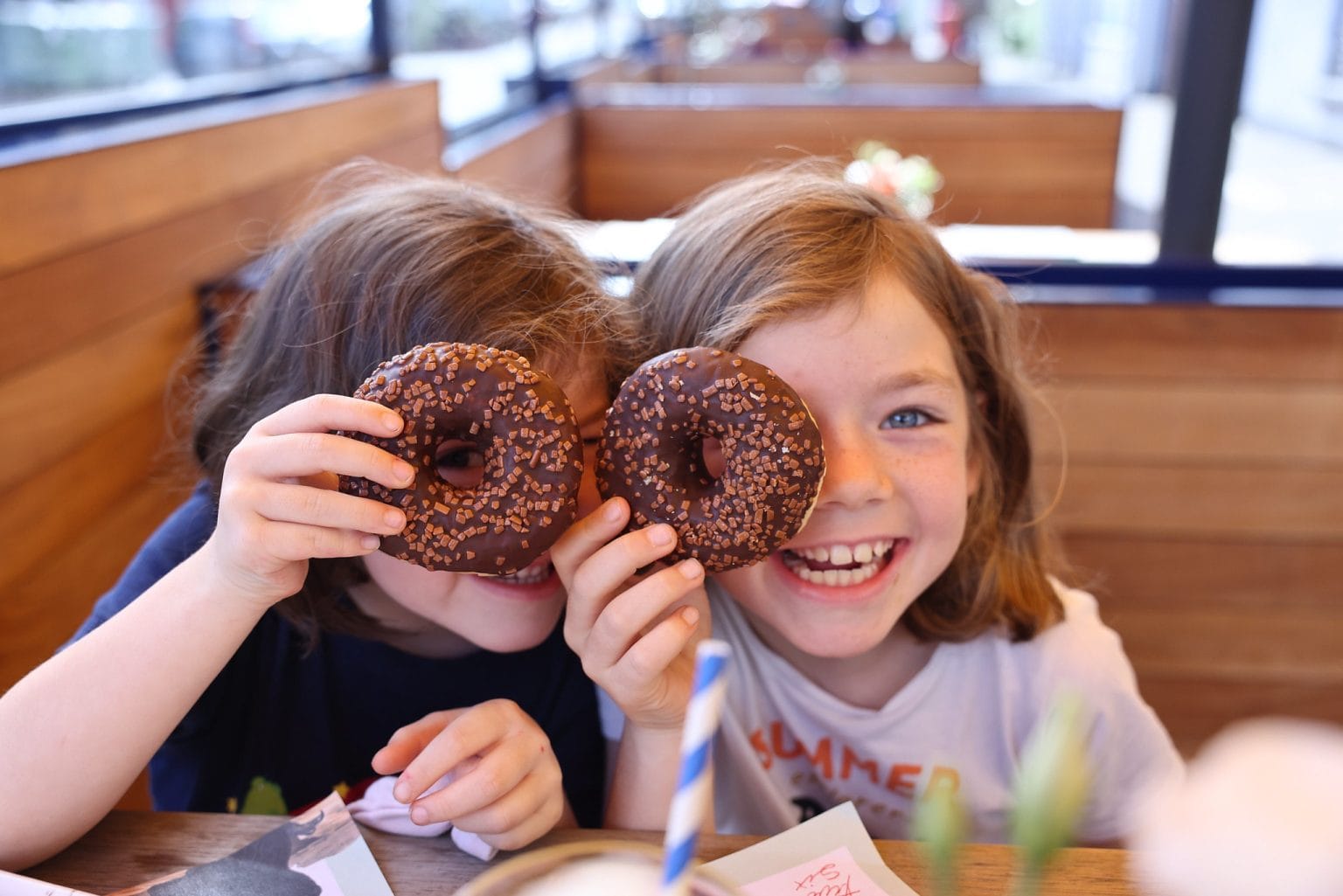 Two young children sitting inside a wooden-paneled cafe, smiling and playfully holding chocolate-covered donuts with sprinkles over their eyes like glasses.