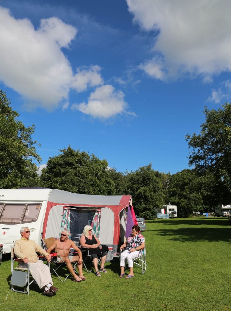 Four people sitting and chatting in camping chairs outside a white and red caravan on a sunny day, with a green grassy field and trees in the background under a partly cloudy blue sky.