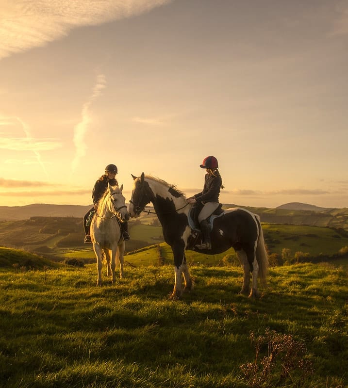 Two people wearing helmets are riding horses on a grassy hill during sunset, with a scenic view of rolling hills and a partly cloudy sky in the background.
