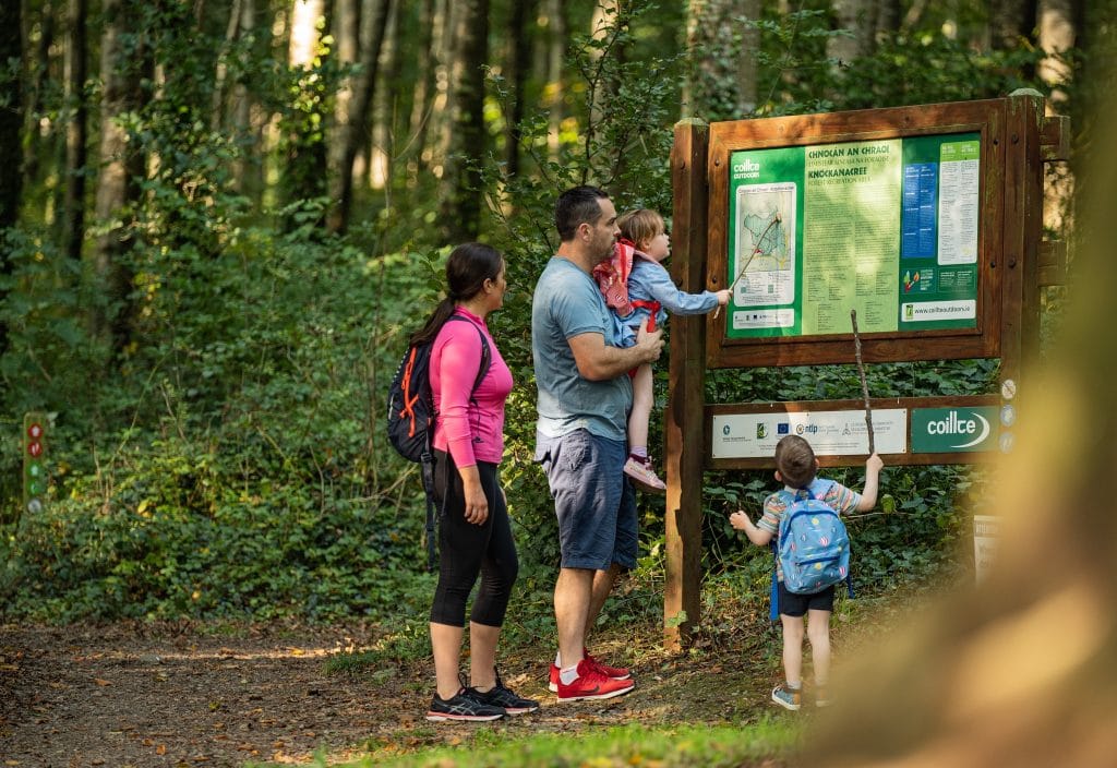 Family of four looking at a wooden information board in a forested hiking area, with two children holding sticks and wearing backpacks.