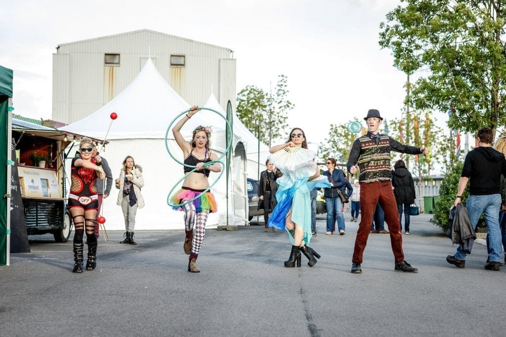 Four street performers dressed in colorful, eclectic costumes are entertaining outdoors near a large white tent. One person is spinning fire sticks, another is playing with multiple hula hoops, a third is dancing with fans and flowing fabric, and the fourth is spinning a diabolo. Several onlookers walk nearby, and there are green trees and industrial buildings in the background.