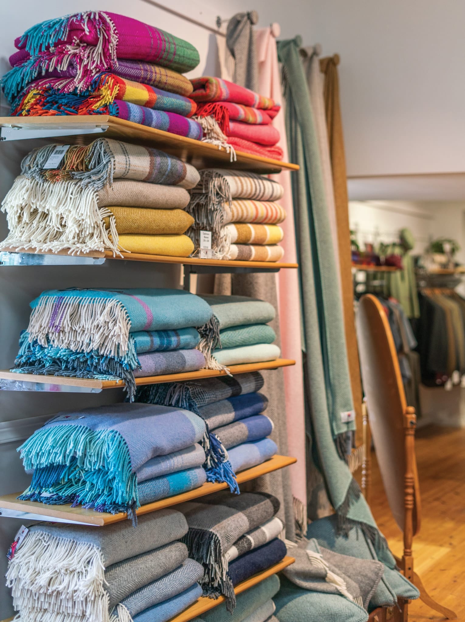 Shelves filled with neatly folded colorful woolen blankets with fringed edges in a cozy fabric store interior, with hanging textiles and clothing racks in the background.