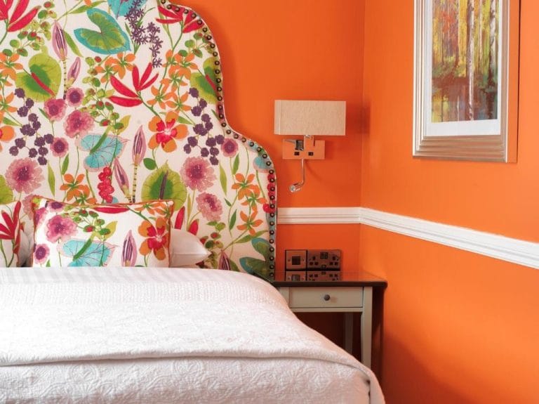 A close-up of a cozy bedroom corner with an orange wall featuring white trim, a colorful floral-patterned upholstered headboard and matching pillow, a white quilted bedspread, a nearby bedside table with a built-in power outlet, a wall-mounted beige lamp, and a framed abstract painting.