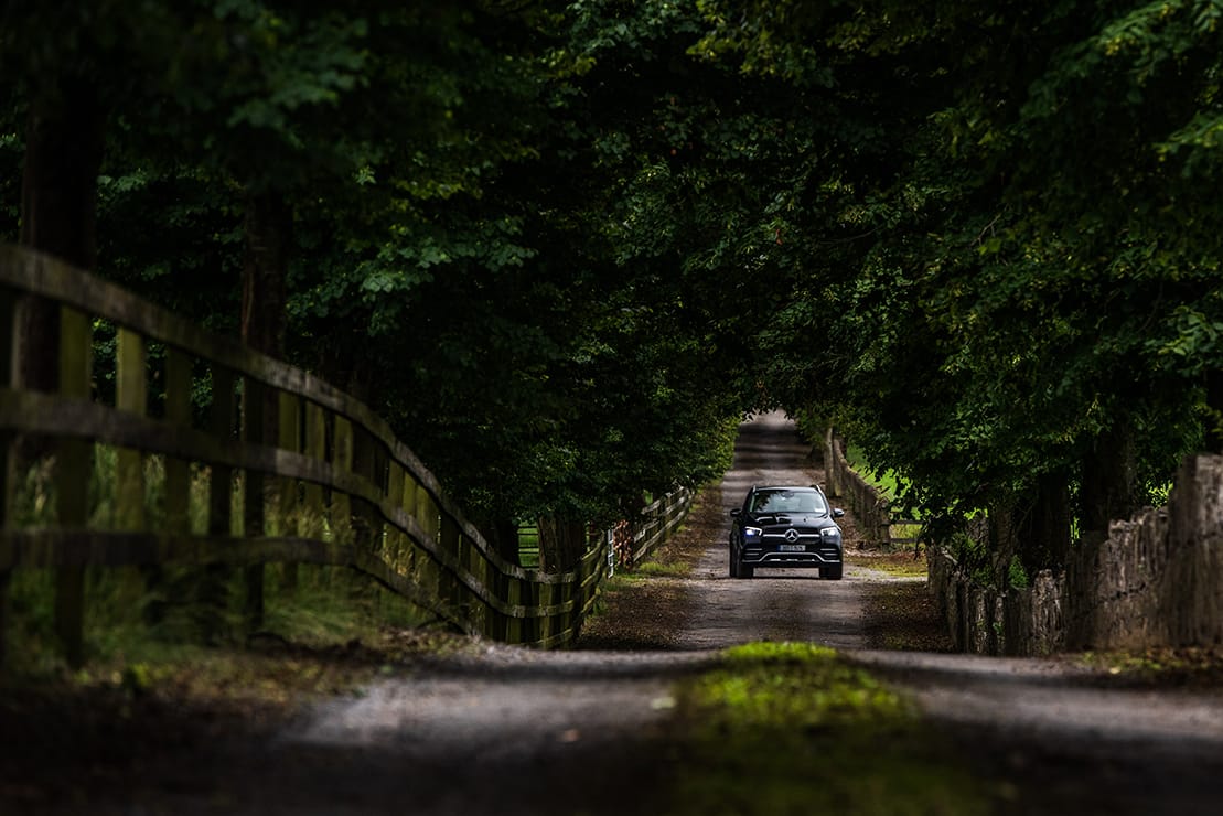 Black Mercedes-Benz SUV driving on a narrow, tree-lined country road with wooden fences on both sides.