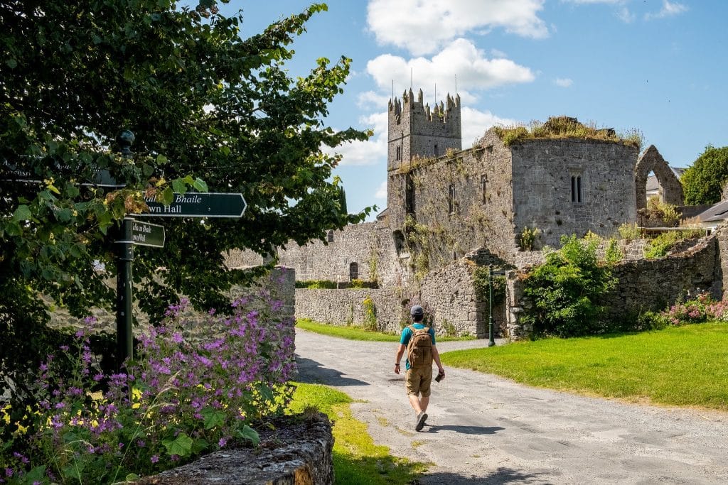 A person wearing a blue shirt, brown shorts, and a hat is walking on a path towards an ancient stone castle with a prominent tower. There are green trees, purple flowers, and a directional signpost along the path under a partly cloudy blue sky.
