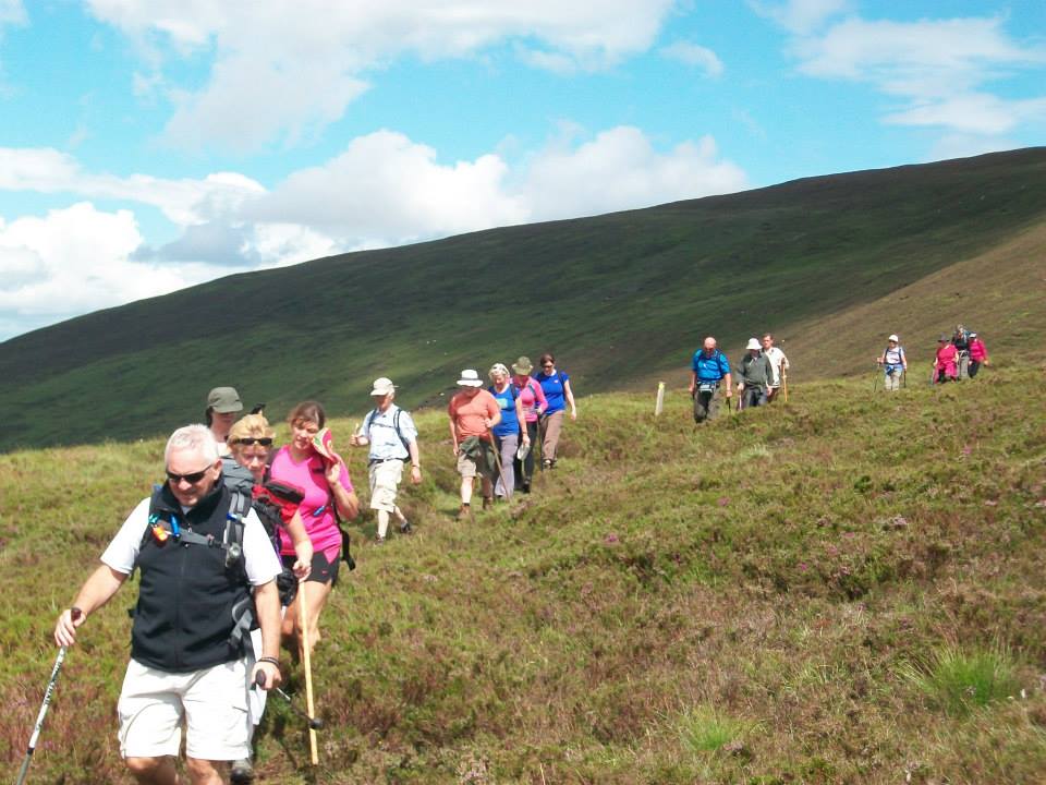 Group of hikers walking in a line on a grassy hillside under a partly cloudy blue sky with a large hill in the background.
