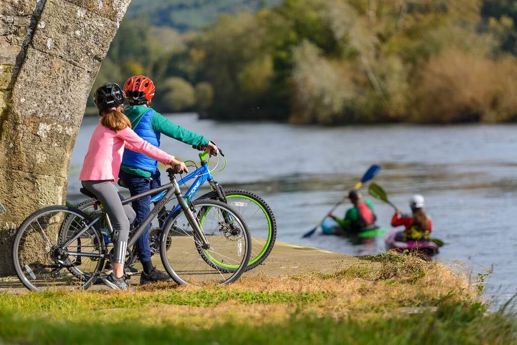 Two children wearing helmets with bicycles stand near a stone arch by a riverside, watching two kayakers paddling on the water.