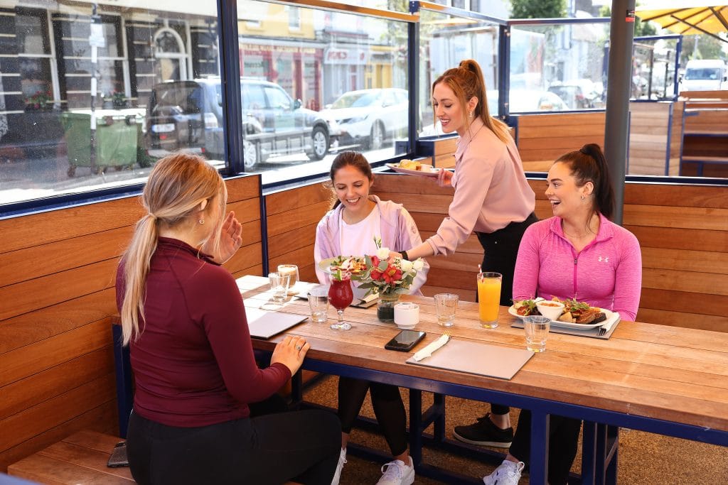 Three women in athletic wear sitting at a wooden table in an outdoor café, smiling and receiving food from a waitress. The table has drinks, plates of food, a smartphone, and a small floral arrangement.