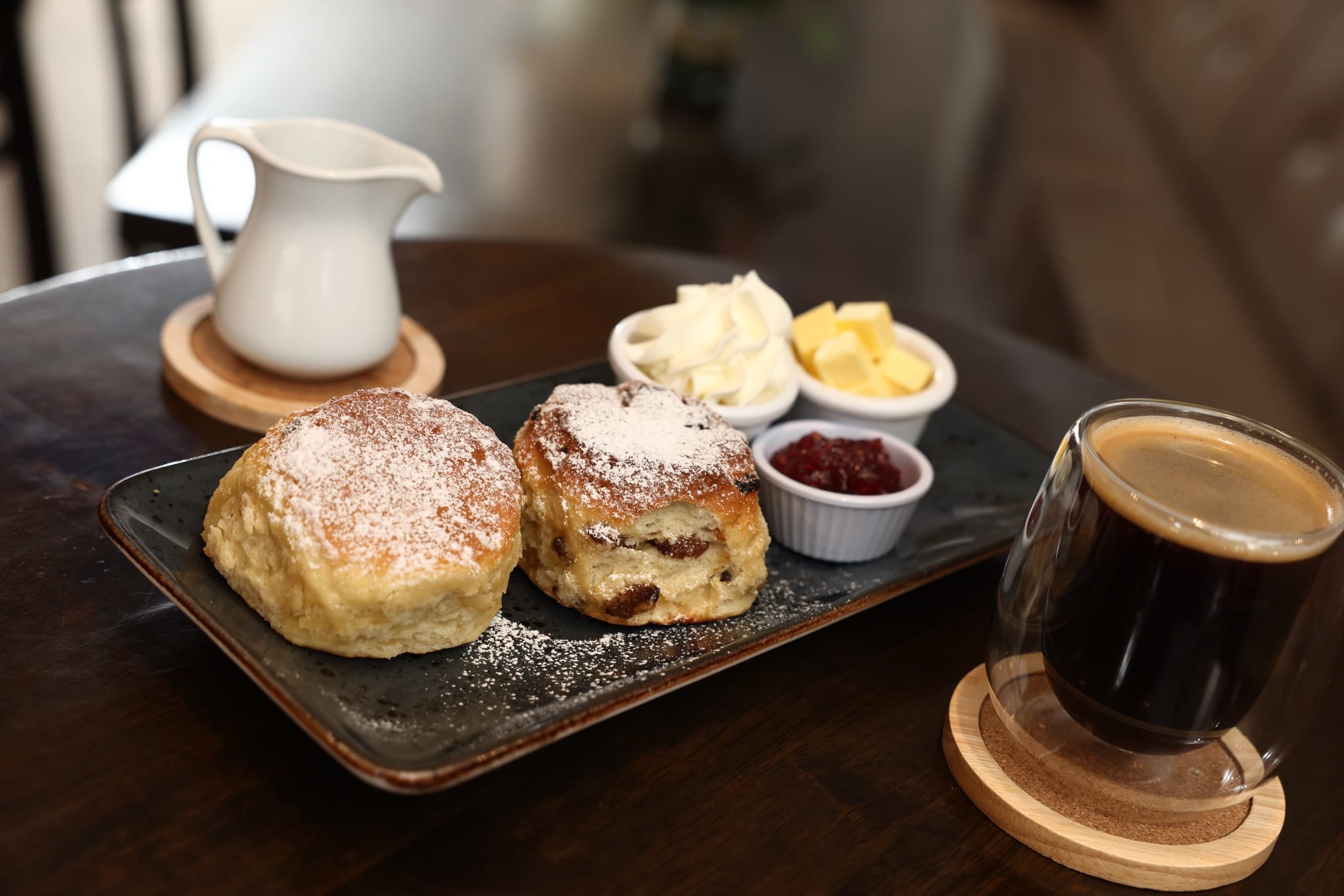 Two powdered sugar-dusted scones on a rectangular black plate with small bowls of clotted cream, butter, and strawberry jam, accompanied by a glass of black coffee and a white pitcher on a dark wooden table.