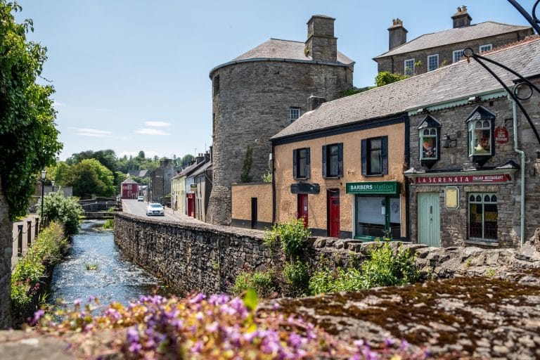 A picturesque street scene along a small river in a quaint town featuring a round stone tower, a beige building with a barbershop, and a stone building housing "La Serenata" Italian restaurant, with purple flowers in the foreground and greenery lining the riverbanks under a clear blue sky.
