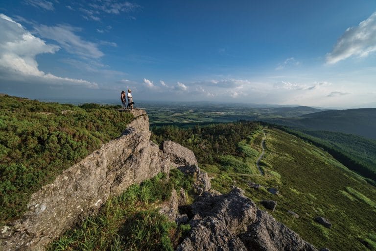 Two hikers stand on a rocky cliff overlooking a vast, green valley with rolling hills under a blue sky with scattered clouds. A winding path runs through the hillside below.