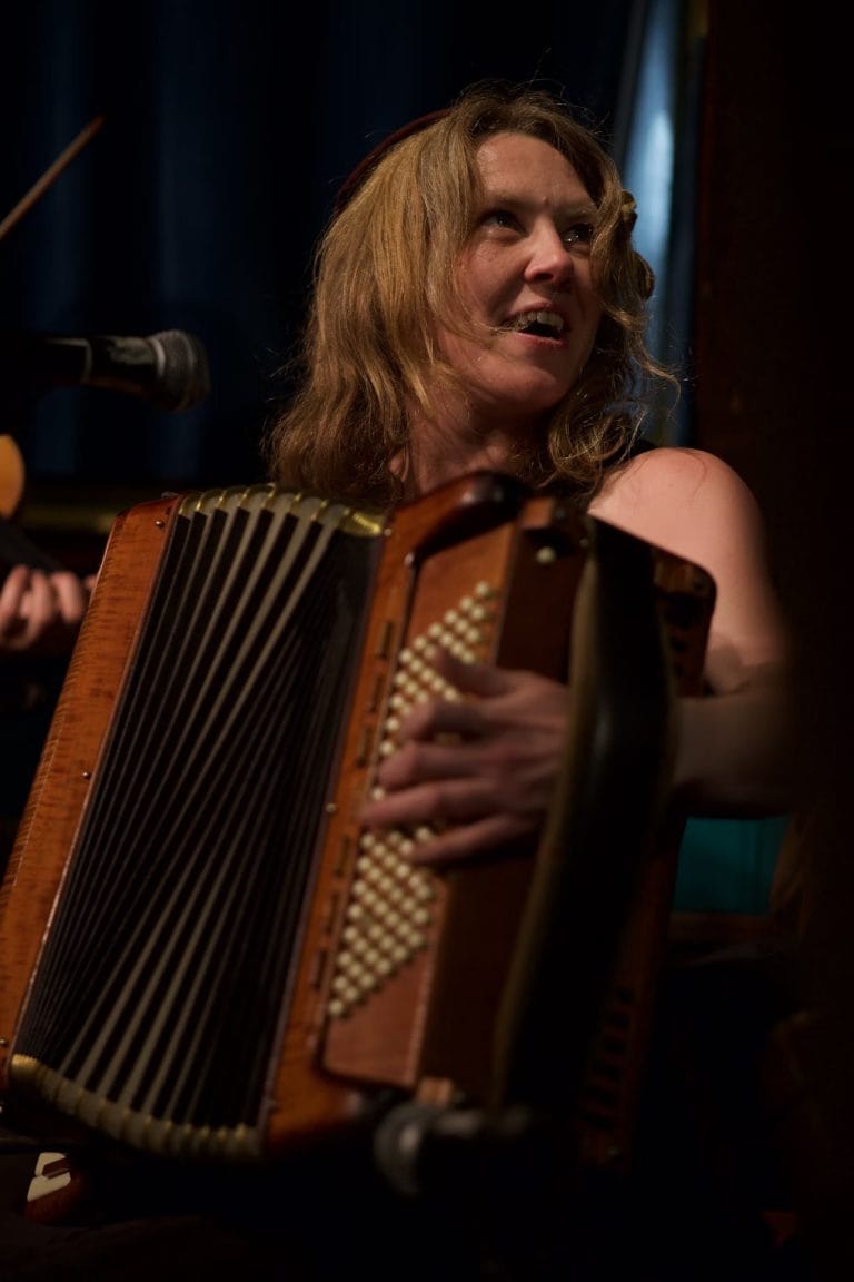 A woman with wavy blonde hair is playing a wooden accordion and looking off to the side, with a microphone in front of her. The background is dark and out of focus.